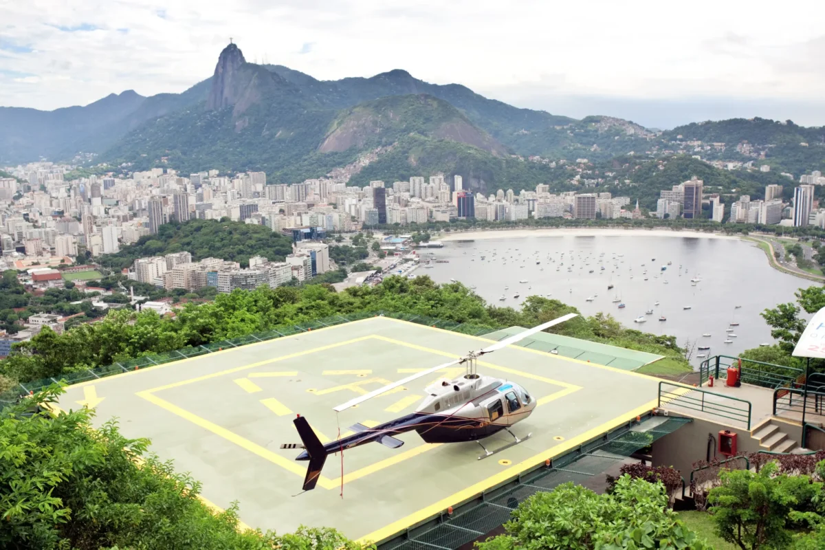 A helicopter is parked on a rooftop helipad against a lush, scenic backdrop and cityscape. Inside, the helicopter pilot protects his aviation license by complying with FAR/AIM regulations